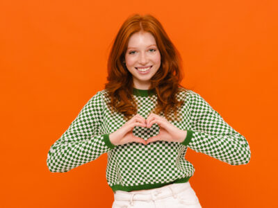 Ginger woman in plaid sweater smiling and showing heart gesture isolated over orange background